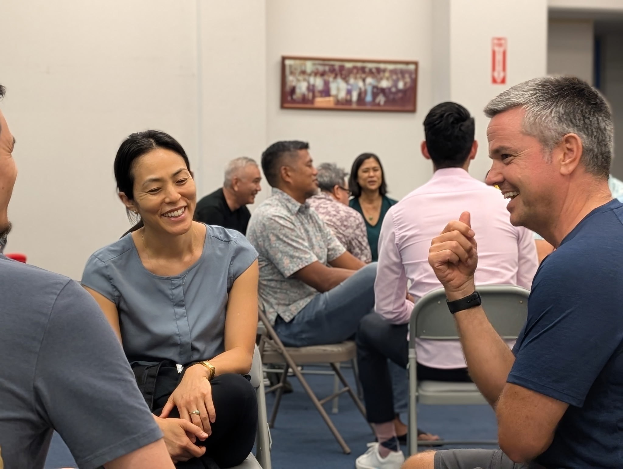 Two colleagues laughing mid-exchange while the rest of the room works in pairs behind them