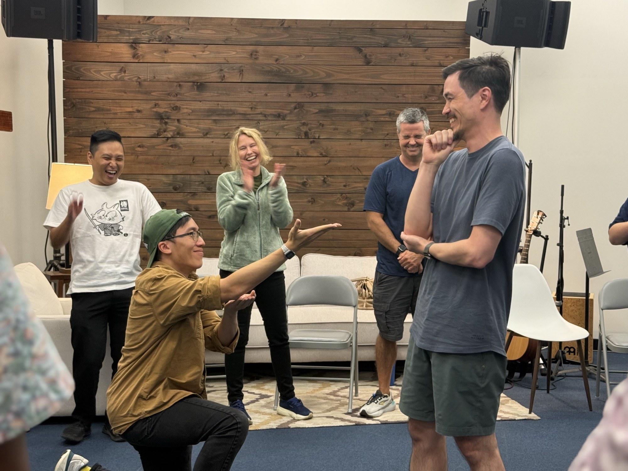 A participant on one knee mid-scene while five colleagues laugh